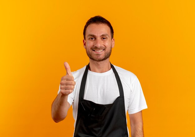 Hombre barbero en delantal mirando al frente sonriendo positivo y feliz mostrando los pulgares para arriba de pie sobre la pared naranja