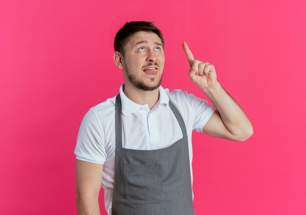 Hombre barbero en delantal mirando hacia arriba apuntando con el dedo con nueva idea de pie sobre fondo rosa