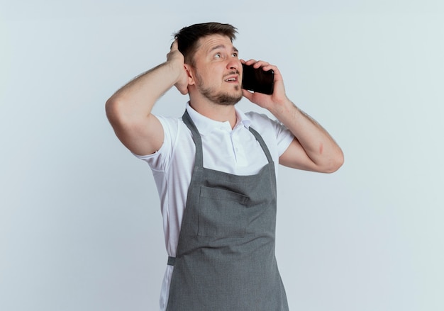 Hombre barbero en delantal mirando confundido mientras habla por teléfono móvil de pie sobre una pared blanca