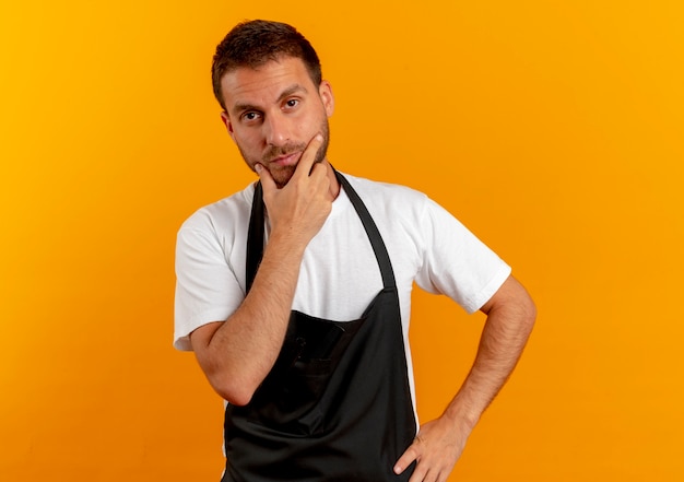 Hombre barbero en delantal mirando hacia el frente con la mano en la barbilla pensando de pie sobre la pared naranja