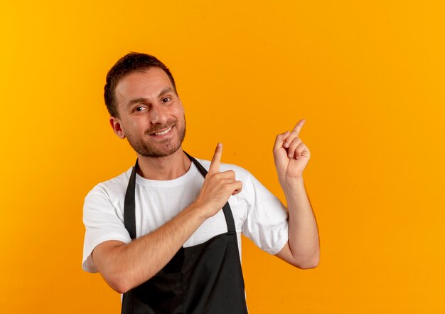 Hombre barbero en delantal mirando hacia el frente sonriendo alegremente apuntando con los dedos hacia el lado de pie sobre la pared naranja