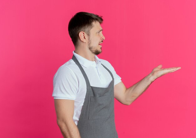 Hombre barbero en delantal mirando a un lado con el brazo extendido como preguntando o discutiendo de pie sobre la pared rosa