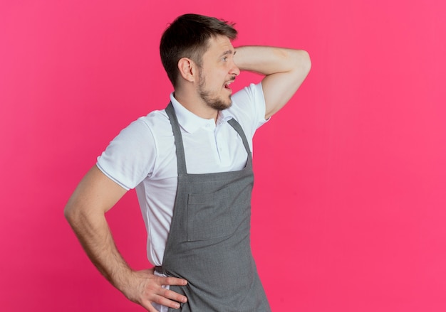 Hombre barbero en delantal mirando a un lado confundido rascarse la cabeza de pie sobre la pared rosa