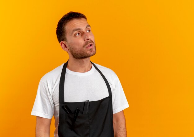 Hombre barbero en delantal mirando a un lado con expresión escéptica de pie sobre la pared naranja