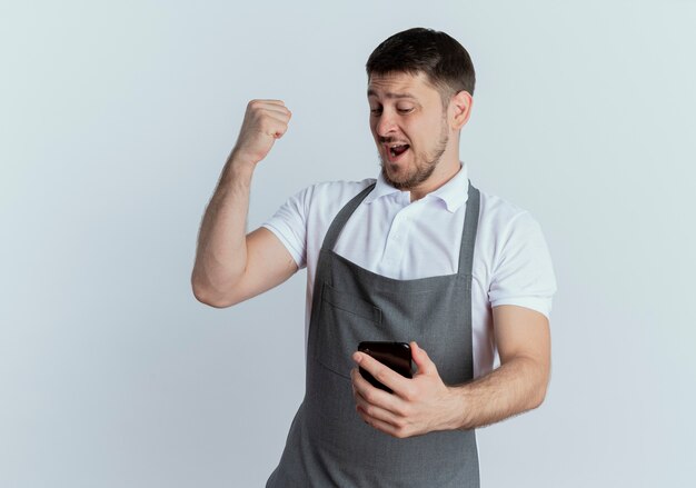 Hombre barbero en delantal mirando la pantalla de su teléfono inteligente apretando el puño feliz y emocionado de pie sobre la pared blanca