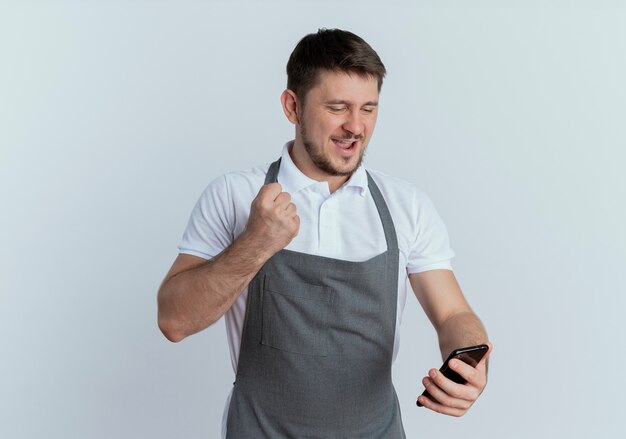 Hombre barbero en delantal mirando la pantalla de su teléfono inteligente apretando el puño feliz y emocionado de pie sobre la pared blanca