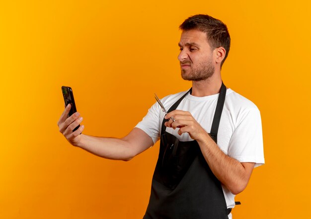 Hombre barbero en delantal mirando la pantalla de su teléfono móvil sosteniendo unas tijeras mirando disgustado de pie sobre la pared naranja