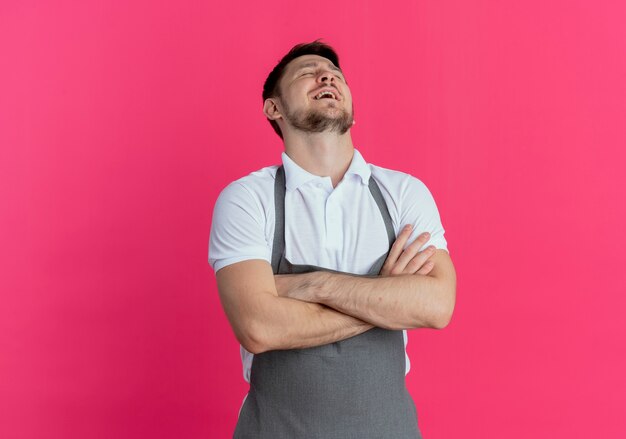 Hombre barbero en delantal molesto y cansado con los brazos cruzados de pie sobre fondo rosa