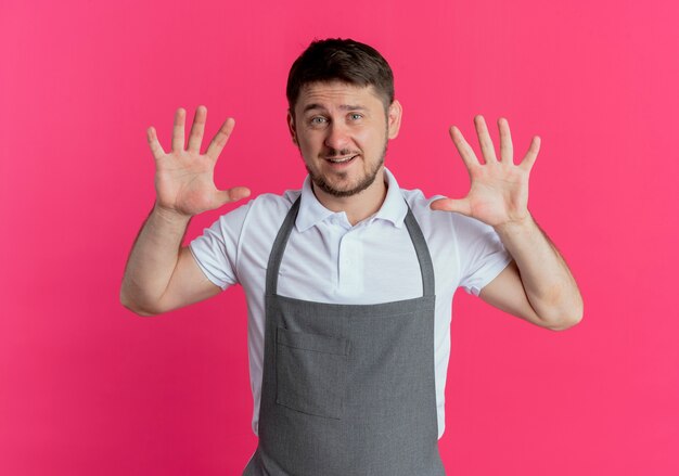 Hombre barbero en delantal mostrando y apuntando hacia arriba con los dedos número diez sonriendo de pie sobre la pared rosa