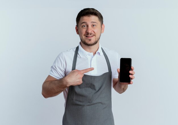 Hombre barbero en delantal mostrando smartphone apuntando con el dedo sonriendo de pie sobre la pared blanca