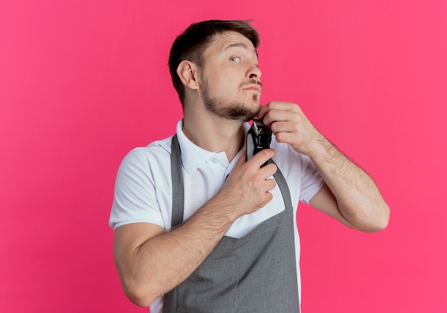 Foto gratuita hombre barbero en delantal recortando su barba de pie sobre una pared rosa