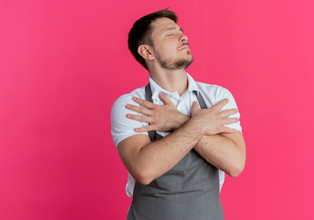 Hombre barbero en delantal sosteniendo los brazos cruzados sobre el pecho sintiéndose agradecido con los ojos cerrados de pie sobre la pared rosa
