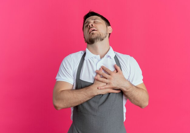 Hombre barbero en delantal sosteniendo los brazos cruzados sobre el pecho sintiéndose agradecido con los ojos cerrados de pie sobre la pared rosa