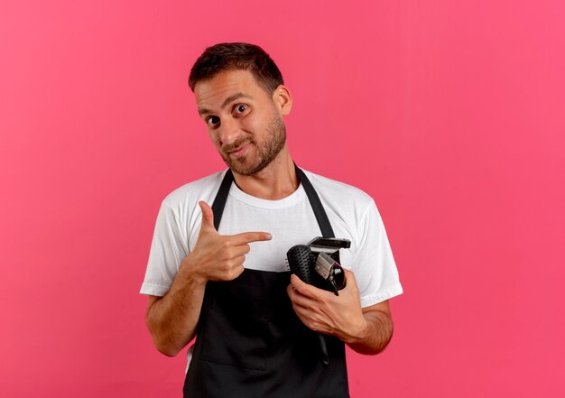 Hombre barbero en delantal sosteniendo cepillo de pelo y recortador apuntando con el dedo sonriendo confiado de pie sobre la pared rosa