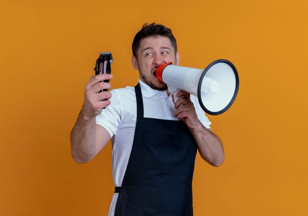 Hombre barbero en delantal sosteniendo recortador de barba gritando al megáfono de pie sobre la pared naranja