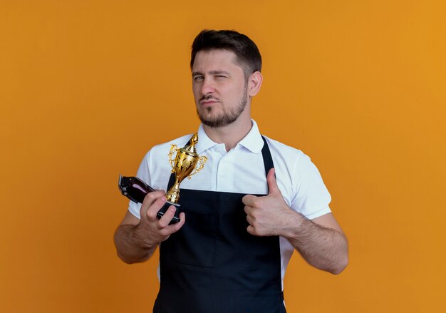 Hombre barbero en delantal sosteniendo recortador de barba y trofeo sonriendo mostrando los pulgares para arriba sonriendo de pie sobre la pared naranja