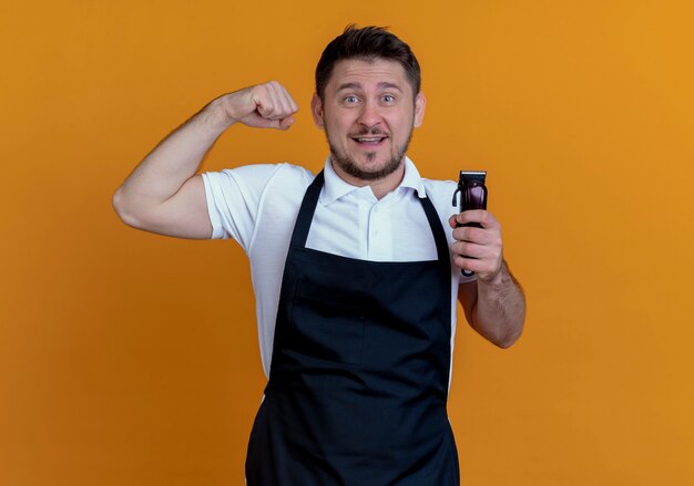 Hombre barbero en delantal sosteniendo recortadora de barba levantando el puño mirando a la cámara sonriendo alegremente de pie sobre fondo naranja