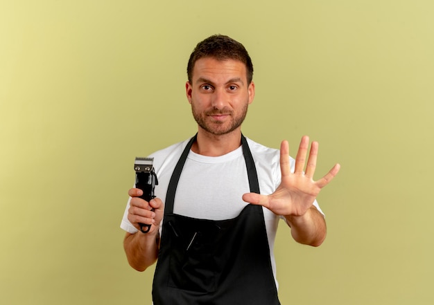 Hombre barbero en delantal sosteniendo recortadora haciendo stop sig con la mano abierta mirando hacia el frente con cara seria de pie sobre una pared de luz