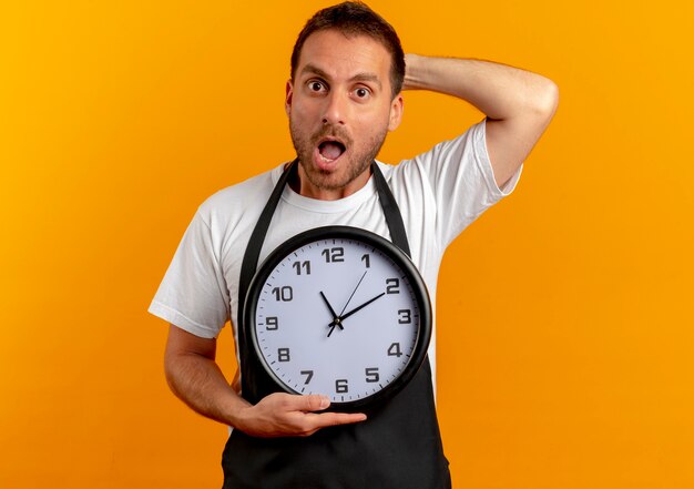 Hombre barbero en delantal sosteniendo reloj de pared mirando hacia el frente preocupado de pie sobre la pared naranja