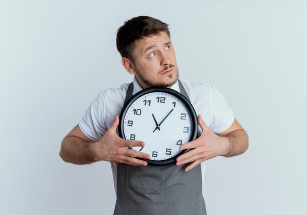 Hombre barbero en delantal sosteniendo reloj de pared mirando a un lado desconcertado de pie sobre la pared blanca