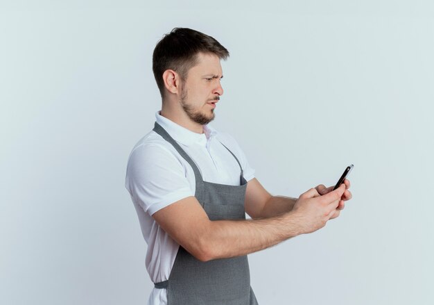 Hombre barbero en delantal sosteniendo smartphone mirando la pantalla con cara seria de pie sobre la pared blanca