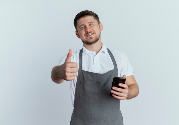 Hombre barbero en delantal sosteniendo smartphone mostrando pulgares arriba sonriendo con cara feliz de pie sobre la pared blanca