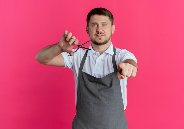 Foto gratuita hombre barbero en delantal sosteniendo tijeras apuntando con el dedo índice de pie sobre la pared rosa