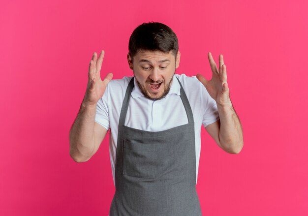 Hombre barbero en delantal volviéndose loco gritando con los brazos levantados de pie sobre fondo rosa