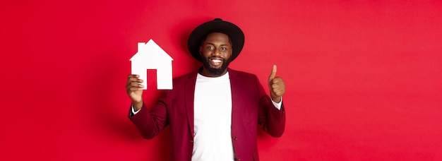 Hombre barbudo afroamericano con estilo de bienes raíces que muestra el pulgar hacia arriba del mercado inmobiliario recomendando la agencia o