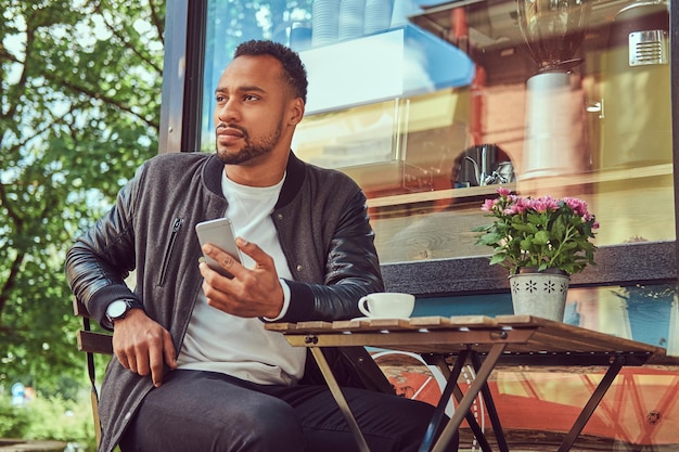 Hombre barbudo afroamericano de moda sentado cerca de una cafetería con una taza de café, usando un teléfono inteligente.