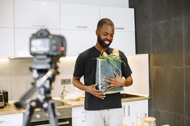 Hombre barbudo afroamericano sonriendo y sosteniendo un paquete con comida. Blogger grabando video para cocinar vlogs en la cocina de casa. Niño con camiseta negra.