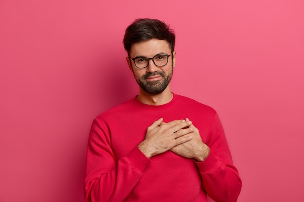 Hombre barbudo agradecido presiona las palmas contra el corazón, siendo conmovido y tocado por palabras agradables, aprecia el regalo recibido, usa anteojos y suéter rosa, expresa gratitud, posa contra la pared rosa