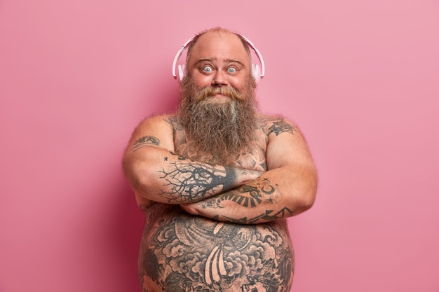 Foto gratuita el hombre barbudo alegre mantiene las manos cruzadas sobre el cuerpo desnudo, se ve feliz, disfruta escuchando música, usa audífonos en las orejas, se ha tatuado el vientre, escucha su canción favorita. ocio en casa, estilo de vida