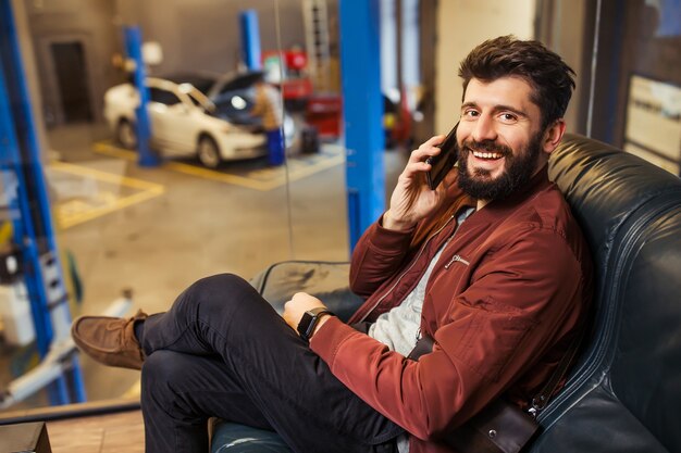 Hombre barbudo alegre sentado en la sala de espera del centro de servicio de automóviles hablando por el teléfono inteligente y mirando a la cámara