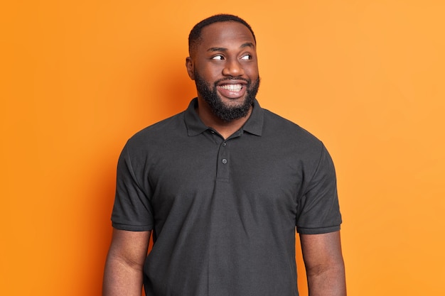 Hombre barbudo alegre sonríe con dientes tiene dientes blancos perfectos mira a un lado con interés viste camiseta negra casual posa contra la pared naranja vívida