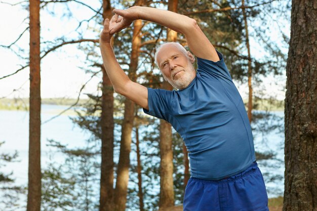 Hombre barbudo anciano autodeterminado enérgico en ropa deportiva posando al aire libre con el bosque y el río, manteniendo los brazos en alto, haciendo curvas laterales, calentando antes de ejecutar el ejercicio