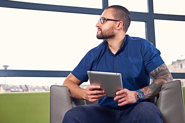 Un hombre barbudo con anteojos y un tatuaje en el brazo se sienta en una silla y usa una tableta.