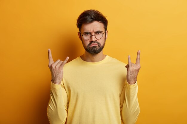 El hombre barbudo de aspecto serio muestra un gesto genial de rock n roll, hace un letrero de heavy metal, es un rockero real, usa gafas ópticas y posa en un puente contra la pared amarilla asiste al concierto de su banda favorita