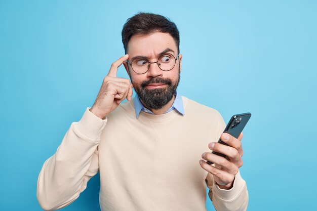 Hombre barbudo avergonzado enfocado en el teléfono inteligente mantiene el dedo en la sien intenta concentrarse en la información mira fijamente la pantalla vestido con poses de suéter limpio contra la pared azul. Concepto de tecnología