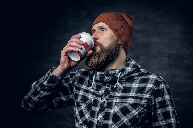 Un hombre barbudo brutal vestido con un sombrero y una camisa de lana, bebe café de un vaso de papel.