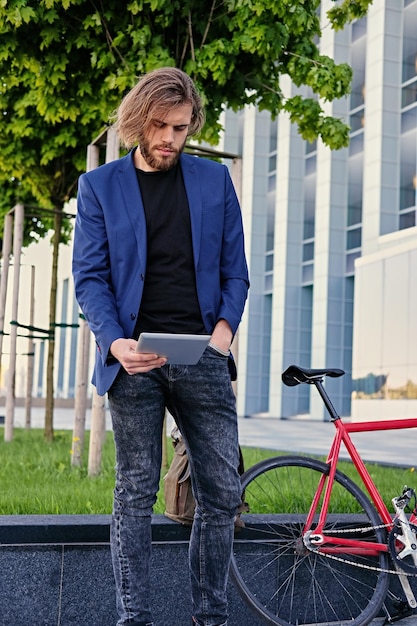 hombre barbudo con cabello largo y rubio sostiene tablet PC con bicicleta roja de una sola velocidad en un parque en el fondo.