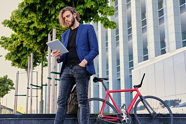 hombre barbudo con cabello largo y rubio sostiene tablet PC con bicicleta roja de una sola velocidad en un parque en el fondo.