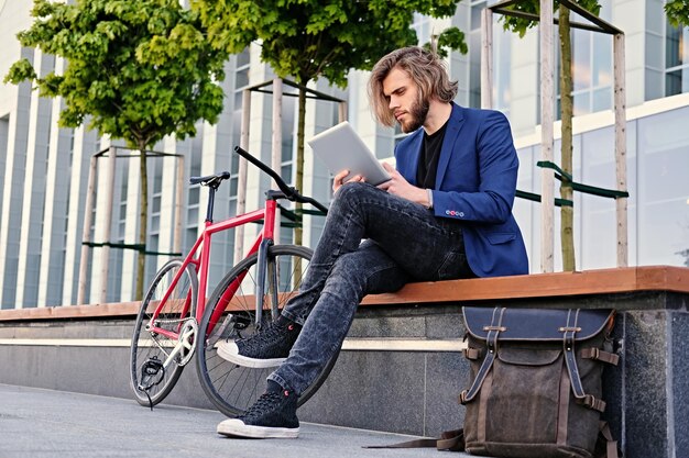 hombre barbudo con cabello largo y rubio sostiene tablet PC con bicicleta roja de una sola velocidad en un parque en el fondo.