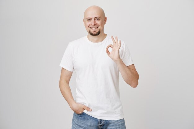 Hombre barbudo calvo satisfecho en camiseta blanca muestra bien firmar, aprobar