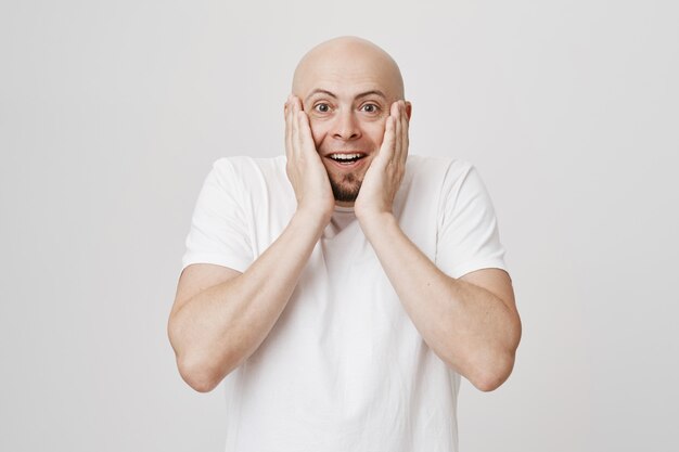 Hombre barbudo calvo sorprendido y feliz toque las mejillas y sonriendo asombrado