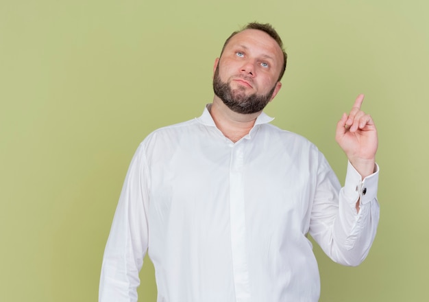 Hombre barbudo con camisa blanca mirando hacia arriba mostrando el dedo índice con cara seria de pie sobre la pared de luz