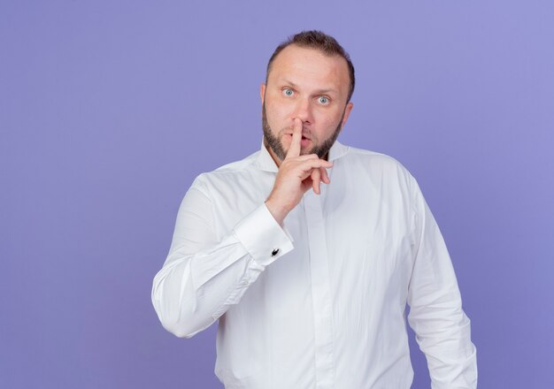 Hombre barbudo con camisa blanca mirando haciendo gesto de silencio con el dedo en los labios de pie sobre la pared azul