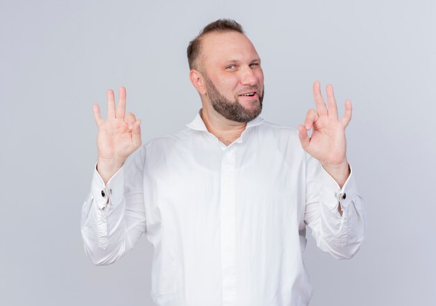 Hombre barbudo con camisa blanca sonriendo confiado mostrando signo ok de pie sobre la pared blanca