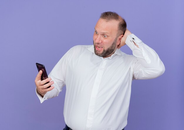 Hombre barbudo con camisa blanca sosteniendo smartphone mirando confundido y sorprendido de pie sobre la pared azul