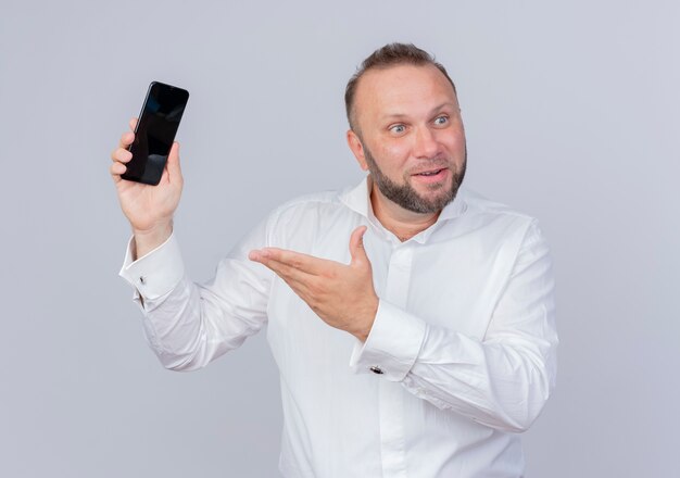 Hombre barbudo con camisa blanca sosteniendo smartphone presentando con brazo feliz y positivo parado sobre pared blanca